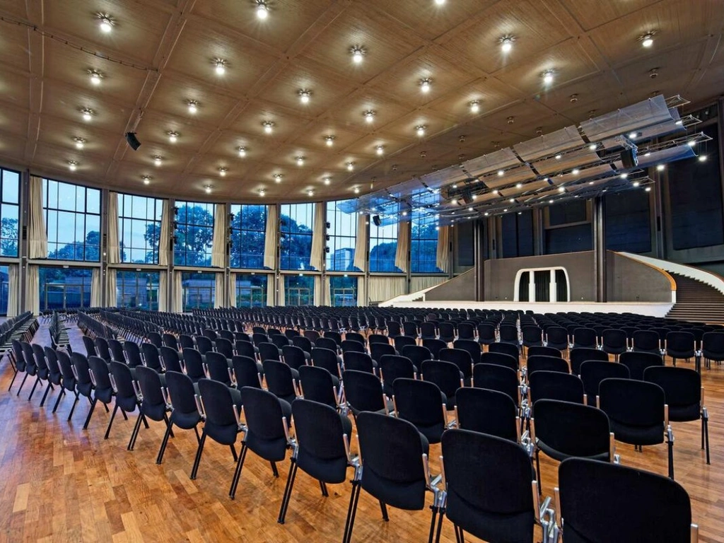 Großer Veranstaltungssaal mit Bühne, vielen Stuhlreihen und hohen Fenstern