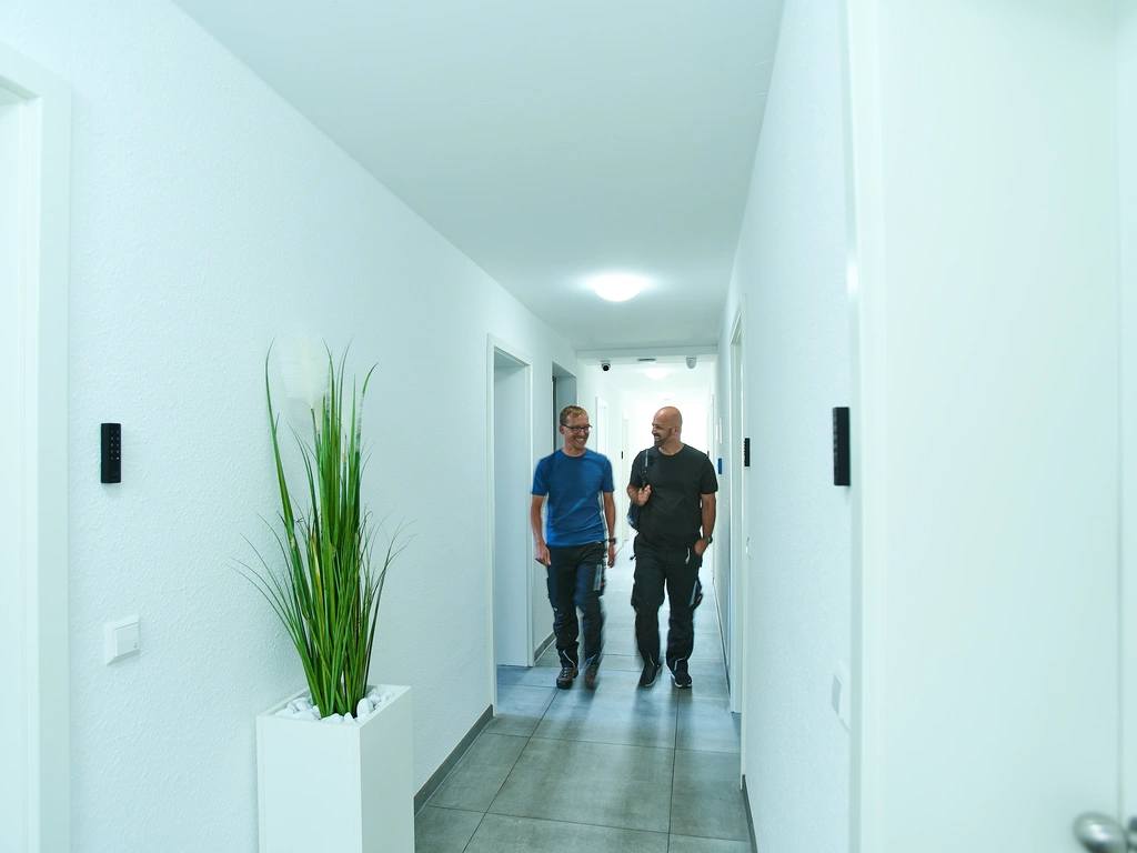 Zwei Männer gehen durch einen hellen Flur mit einer hohen Pflanze